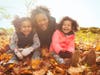 Portrait playful mother and daughters in autumn leaves in sunny park