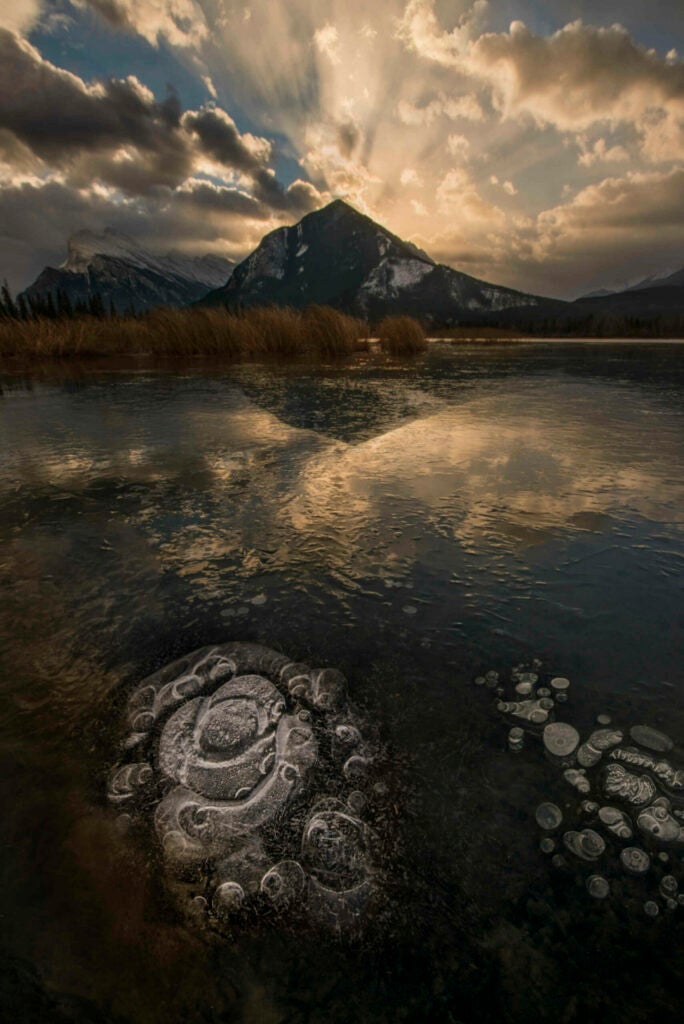 Some landscape scenes can be photographed without much effort, but this particular scene, in Banff National Park took a bit more effort. Sunrise over the Vermillion Lake was stunning but this scene presented 2 problems: a huge depth of field and a huge dynamic range. To make these frozen air bubbles stand out, I had to get very close to them. Mt. Rundle, in the background, was a mile away. The bubbles sat hidden from the sun while the bright sky cast rays out from the mountains. I had to capture several photos, focusing both near and far, and then repeat using various exposures, and then combine in Photoshop to make this photograph.