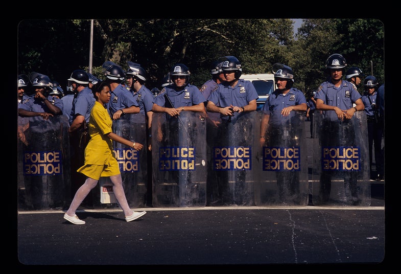 Brooklyn, 1991. A woman walks by a line of police during the Crown Heights race riots in Brooklyn. This was a three-day racial riot that occurred from August 19th to 21st and pitted African American and Caribbean Americans against Jewish residents.