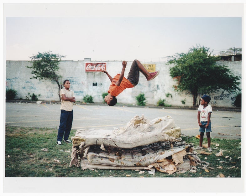 Photograph by Jamal Shabazz. Young kid flipping on matress. This image was made in the Brownsville section of Brooklyn in 1981. Despite conditions of the matress and possible harm, inner city youth improvised and made the best out of what encompassed them.