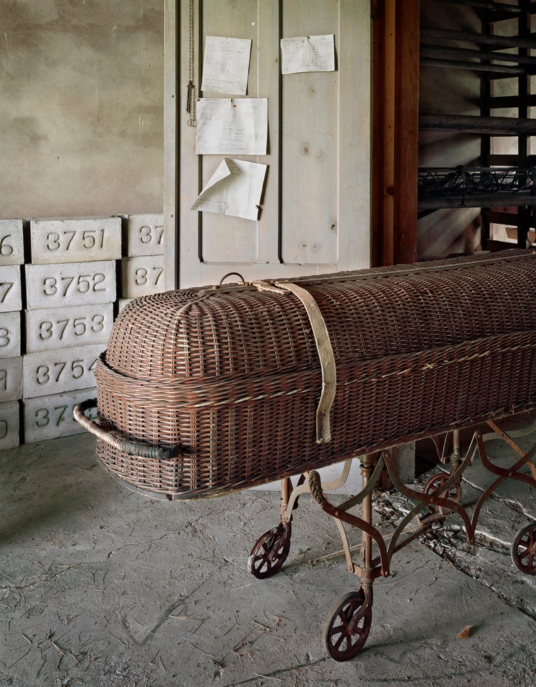 "Hospital staff used this wicker casket to transport bodies from the hospital to the facility’s vast cemetery. This unheated storage room in a cinderblock outbuilding is where the deceased were placed on stretchers during the winter months when the ground froze and the digging of graves was put on hold. In the background are unused grave markers. None of the cemeteries in the institutions that I visited had traditional headstones with patients’ names. Only numbers. The cemetery layouts were often organized by religion: Roman Catholic, Protestant, and Jewish."