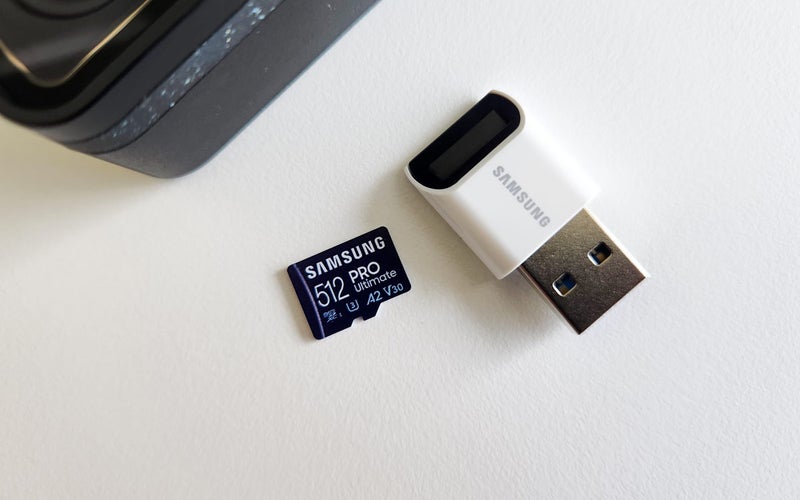Samsung PRO Ultimate 512GB microSDXC memory card for cameras next to a microSDXC card reader and GoPro action camera on a white desk.