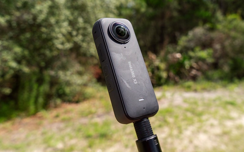 The Insta360 X3 on a selfie stick in front of woods.