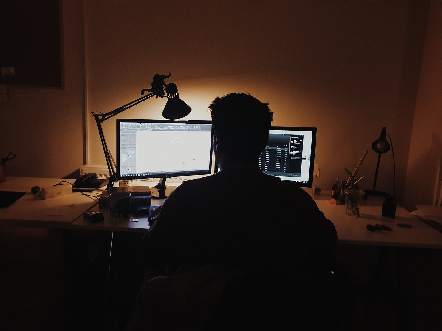 person sitting in a dark room with a lamp and two computer monitors on a desk