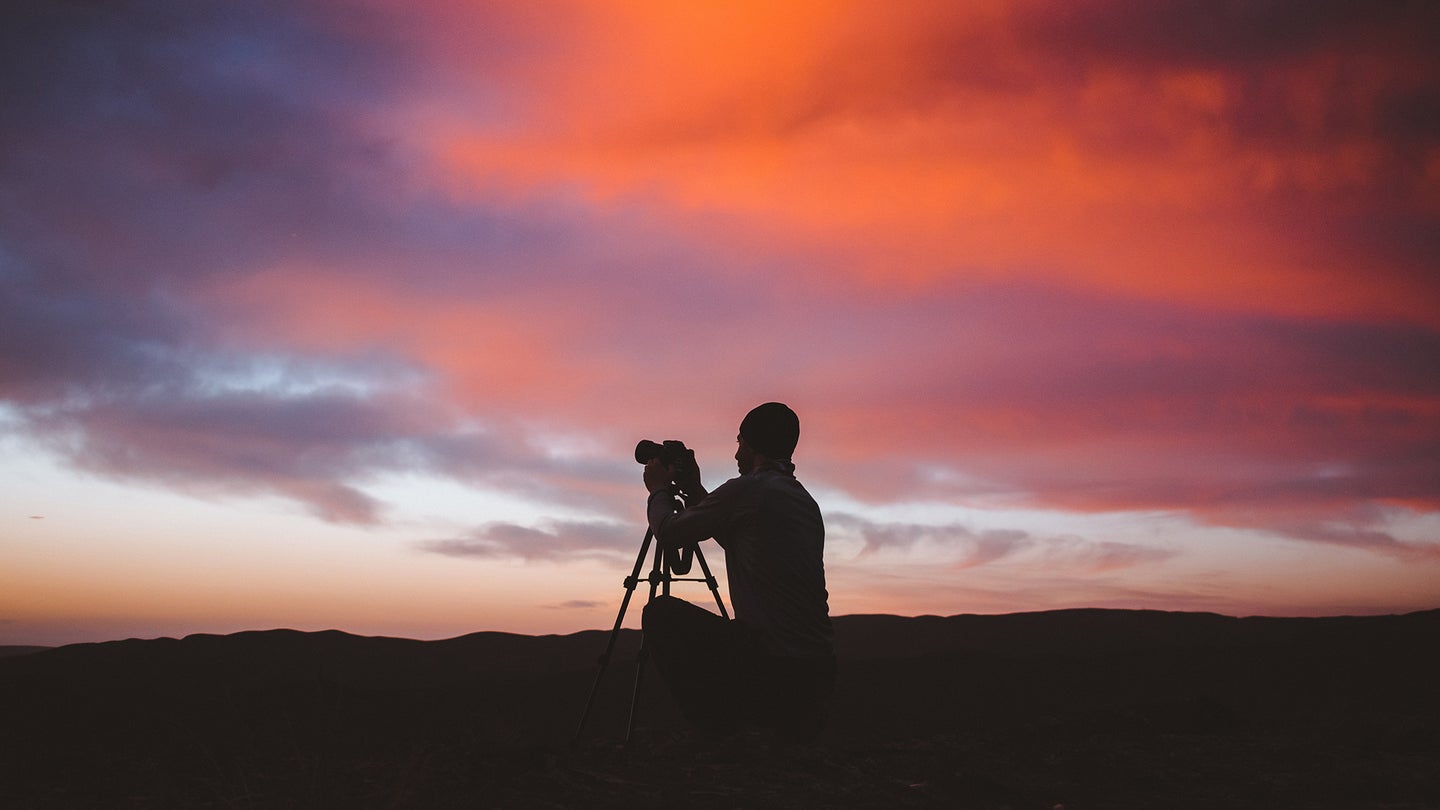 guy with a camera in a tripod with an organge and purple sky behind him