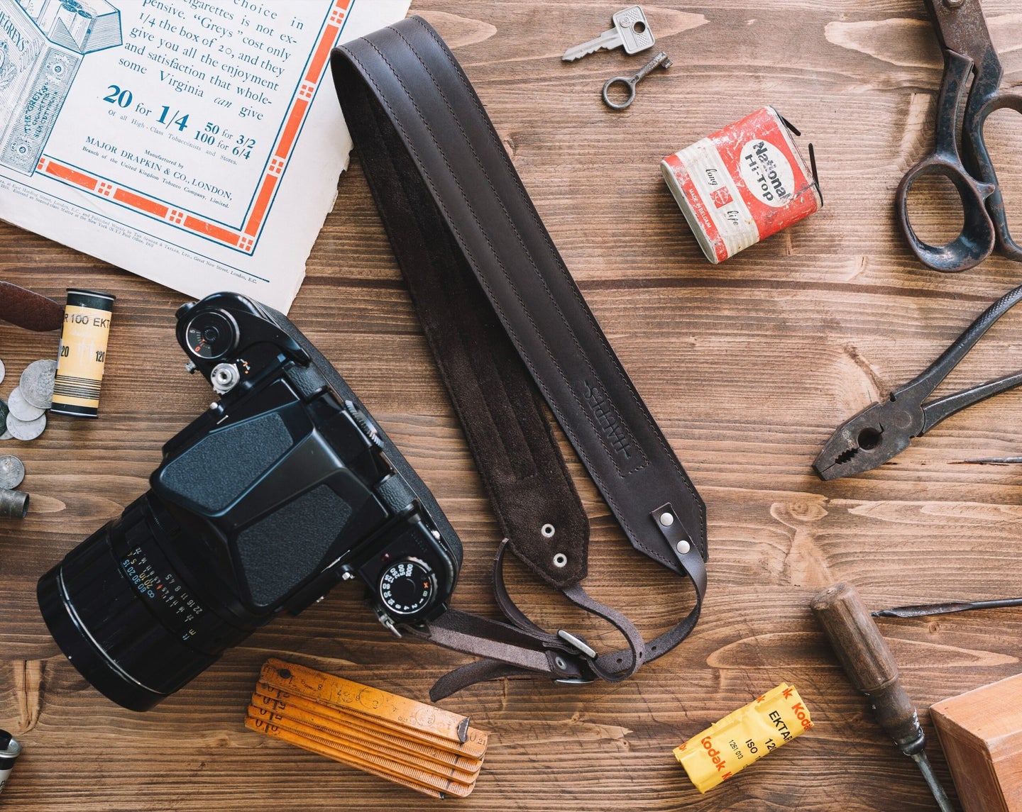 leather camera strap attached to a black camera with the lens, pliers, screwdrivers and meter on a wooden floor