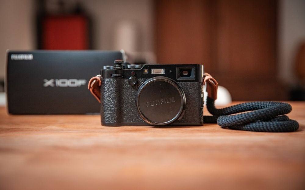 A focused black Fujifilm camera with a neck holder like a black rope resting on a wooden floor with a blurry view from behind.