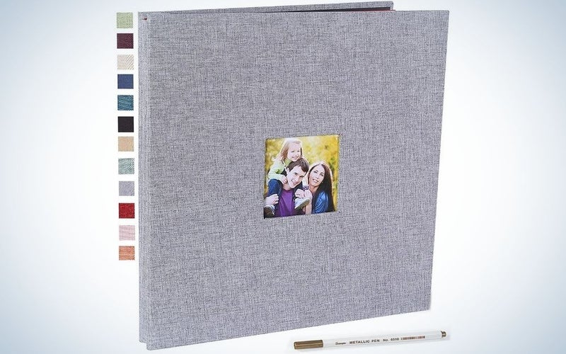 A light grey square photo album with a golden picture of a family in the middle of the album and also a metallic pen in front of it.