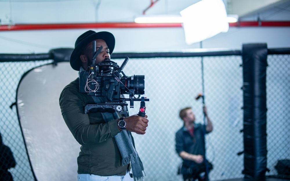 A man in a gray hat and shirt and a scarf who is standing with a professional black camera in his hand.