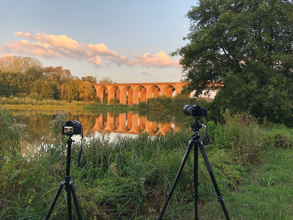A camera set up on the best travel tripod in front of a lake with a bridge spanning it surrounded by greenery.