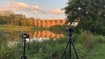 A camera set up on the best travel tripod in front of a lake with a bridge spanning it surrounded by greenery.
