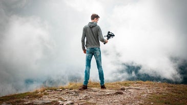 A boy in jeans standing on a rock with a professional camera in his hand filming clouds in front of him.