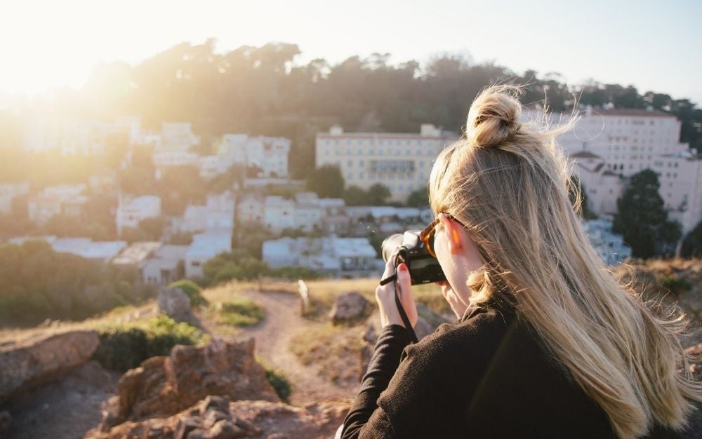 A blonde woman with a black camera in her hands taking pictures in front of the sun and some white houses.