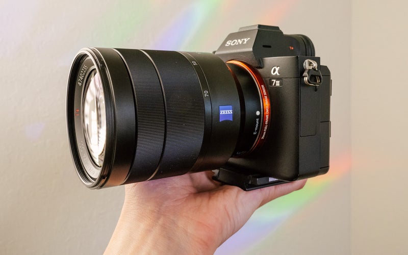 יד מחזיקה את Sony Vario-Tessar T* FE 24-70mm f/4 Lens ואת מצלמת Sony a7 III מול קיר לבן עם קשתות בענן.