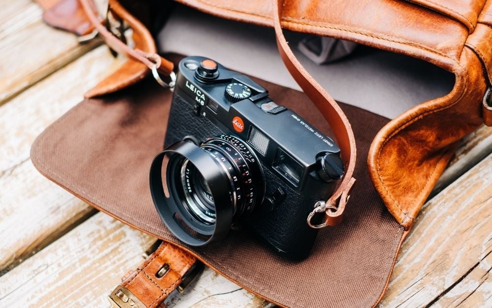 A professional black camera with some orange leather straps placed on the pocket of a brown bag.