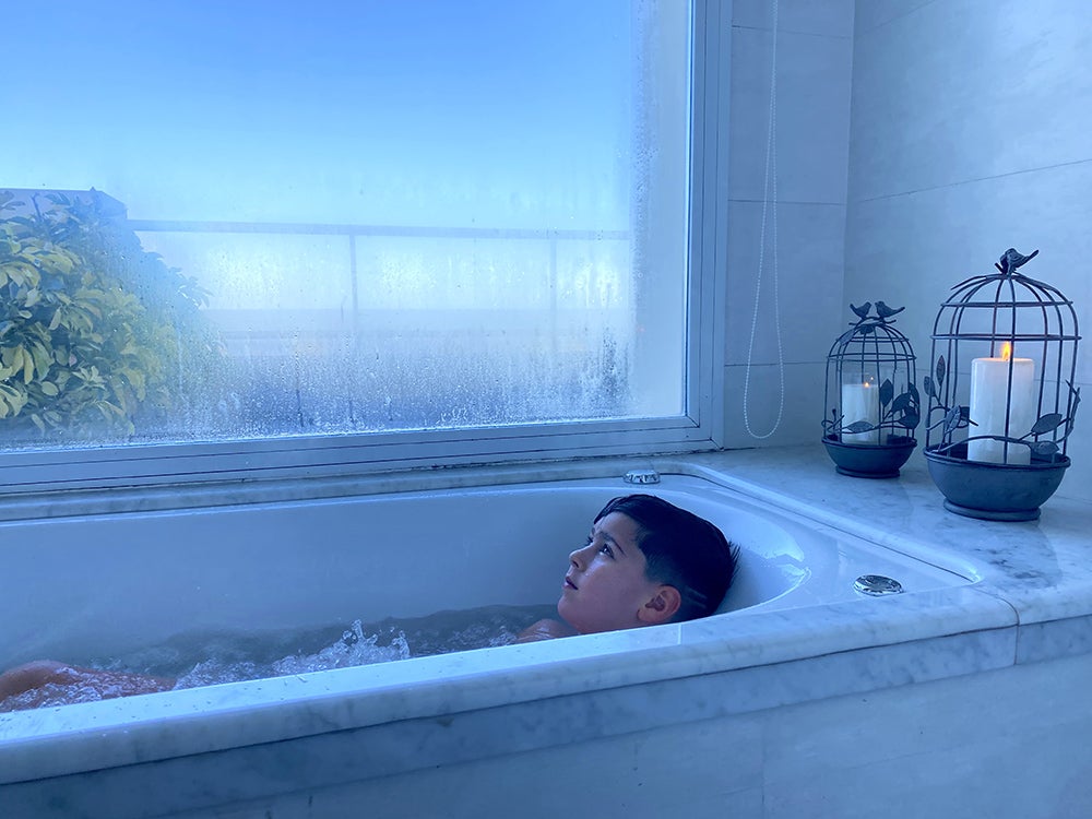 A boy in a tub next to a window streaming in blue light.