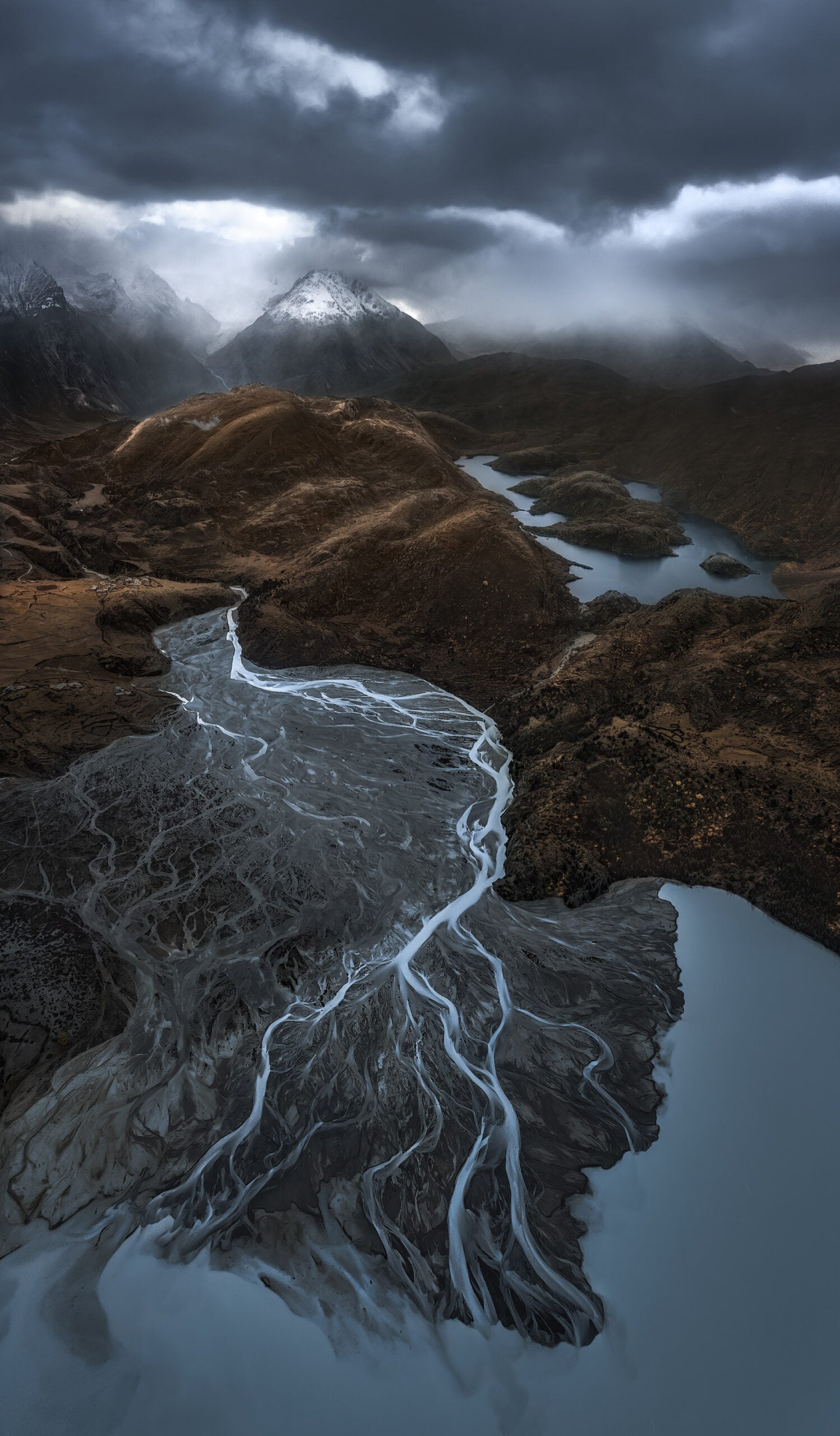 A moody landscape photo with mountains, rivers, streams, and a very cloudy sky.