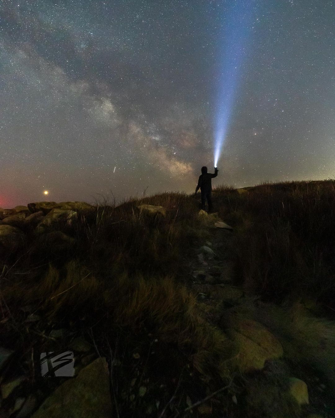 A loan person makes their way through a dark landscape with a flashlight and a sky full of stars.