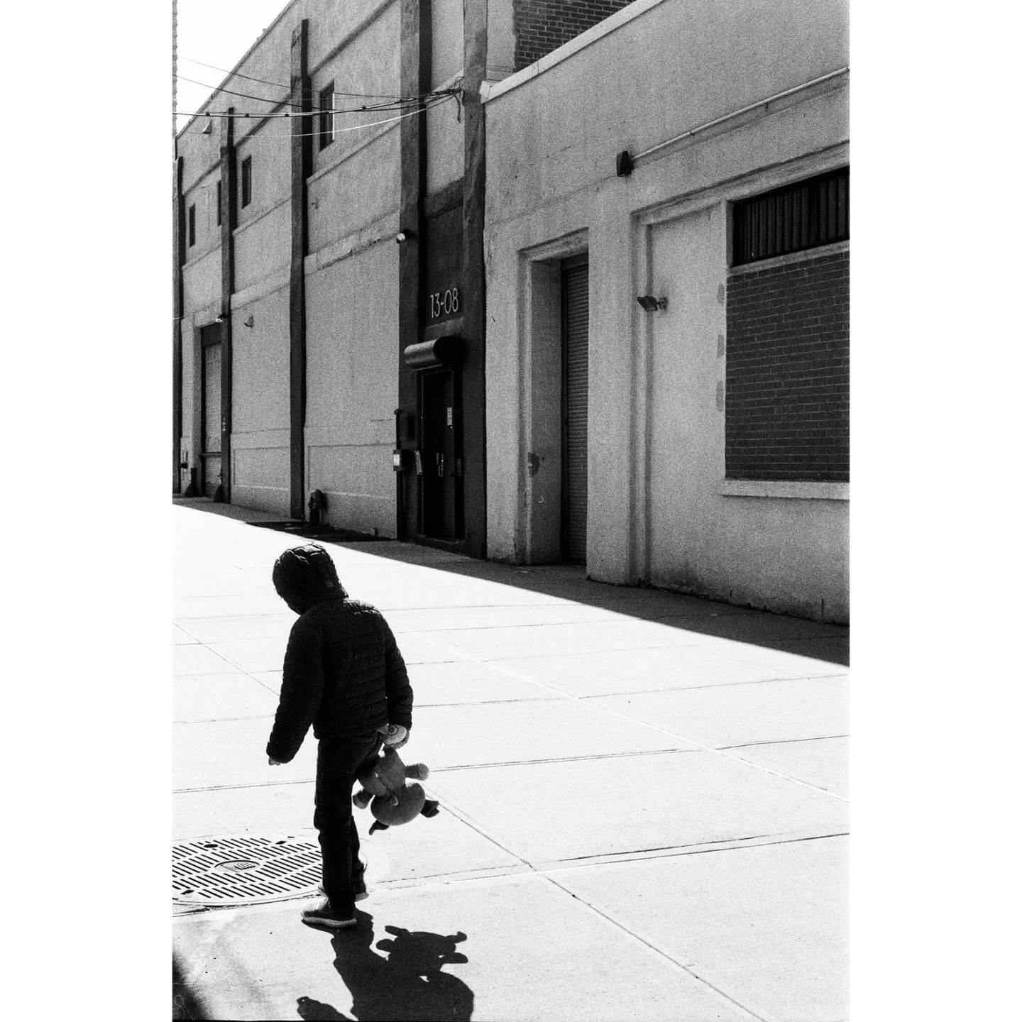 black and white photo of a child holding a stuffed toy walking on the sidewalk. you can only see the child's back and the shadow