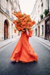 a young woman in a billowing orange dress holds a bunch of orange dahlias in front of her face