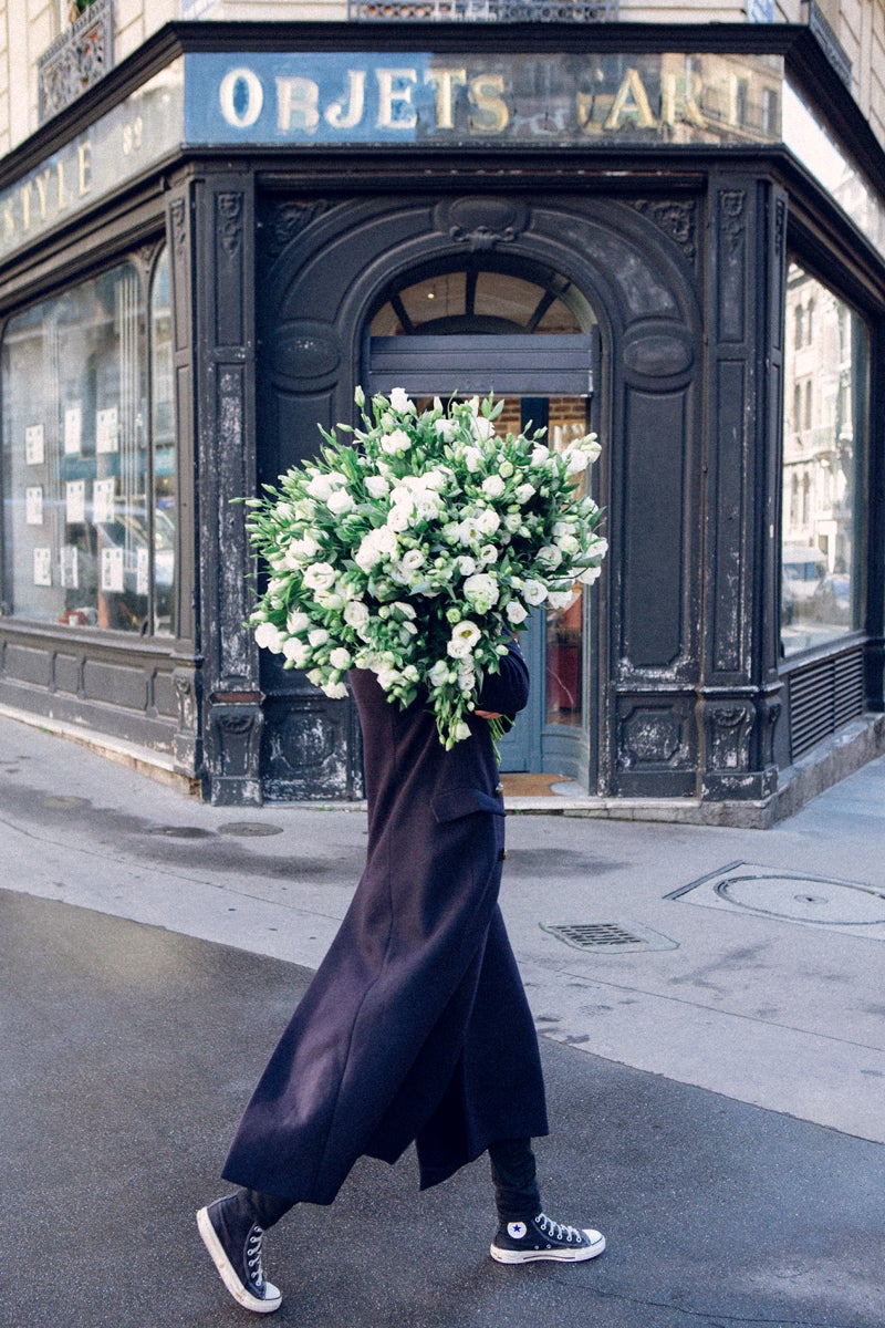 a girl in high tops and a long blue coat walks in paris with an armful of flowers