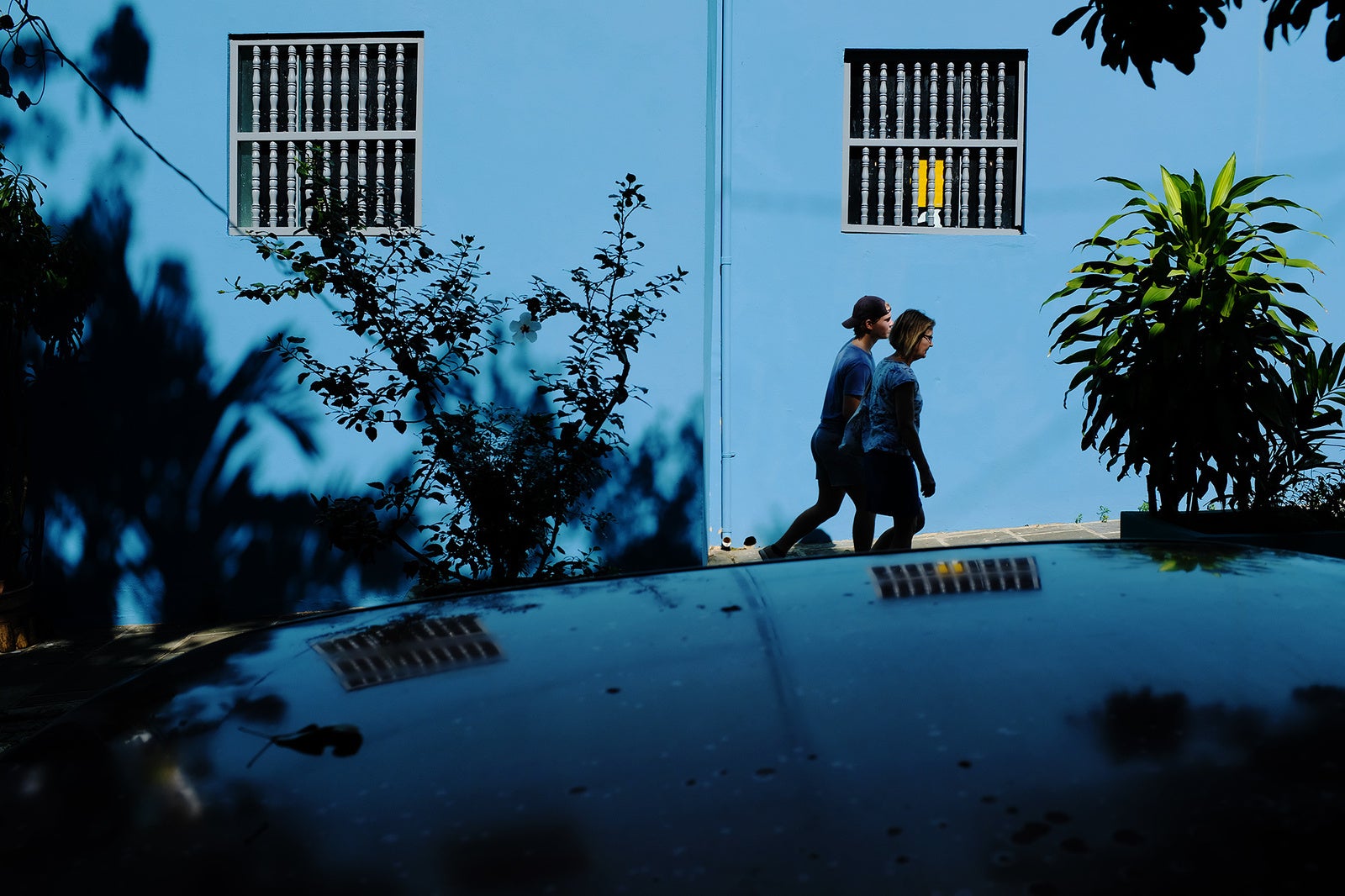 Two people walking next to a bright blue building on a contrasty day.