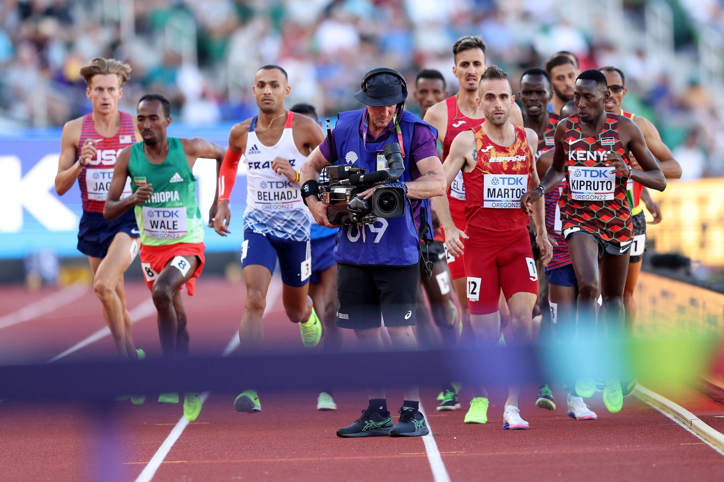 Athletes in the Men's 3000m Steeplechase Final run around a cameraman on the track.
