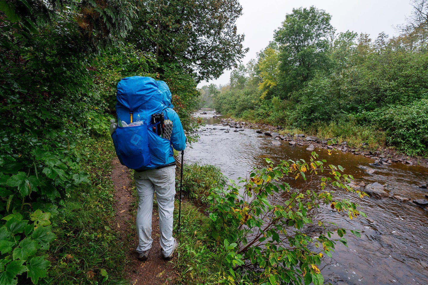A person stands wearing a packed camera backpack on a backpacking trip next to a river