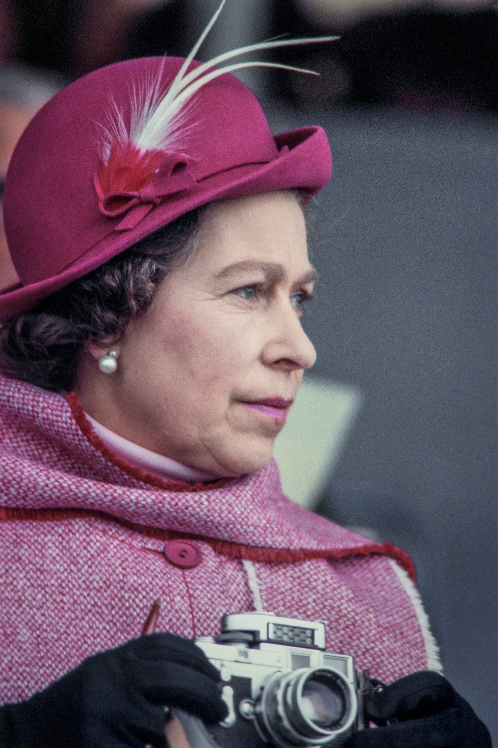 Close-up of British monarch Queen Elizabeth II, in a pale red, feathered, felt hat and a wool cape, as she holds a Leica M3 camera while attending an equestrian event, Windsor, England, circa 1975.