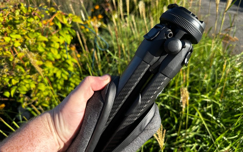 A hand holds the Peak Design Travel Tripod against green foliage.