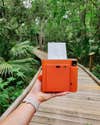 A hand holding the Terracotta Orange Instax Square SQ1 in a green forest.