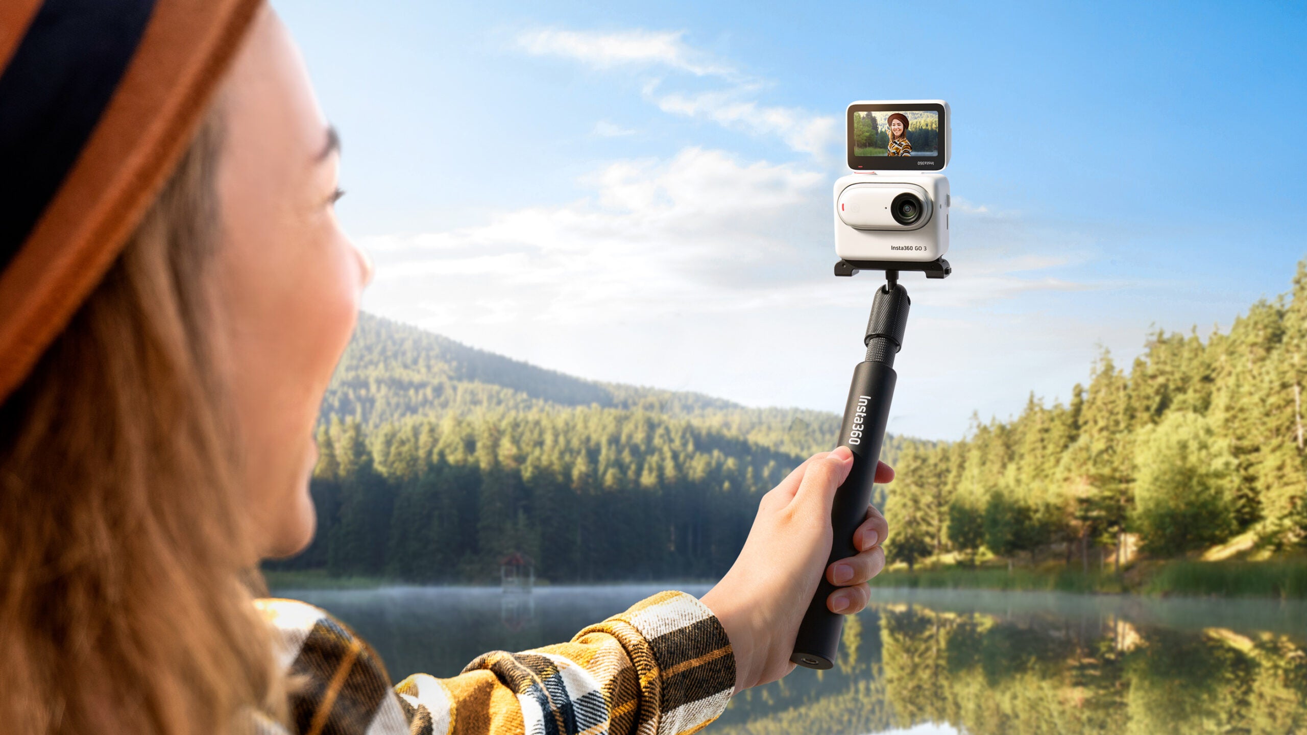 A woman holds the Insta360 GO in the Action Pod on a selfie stick in front of a mountain lake.