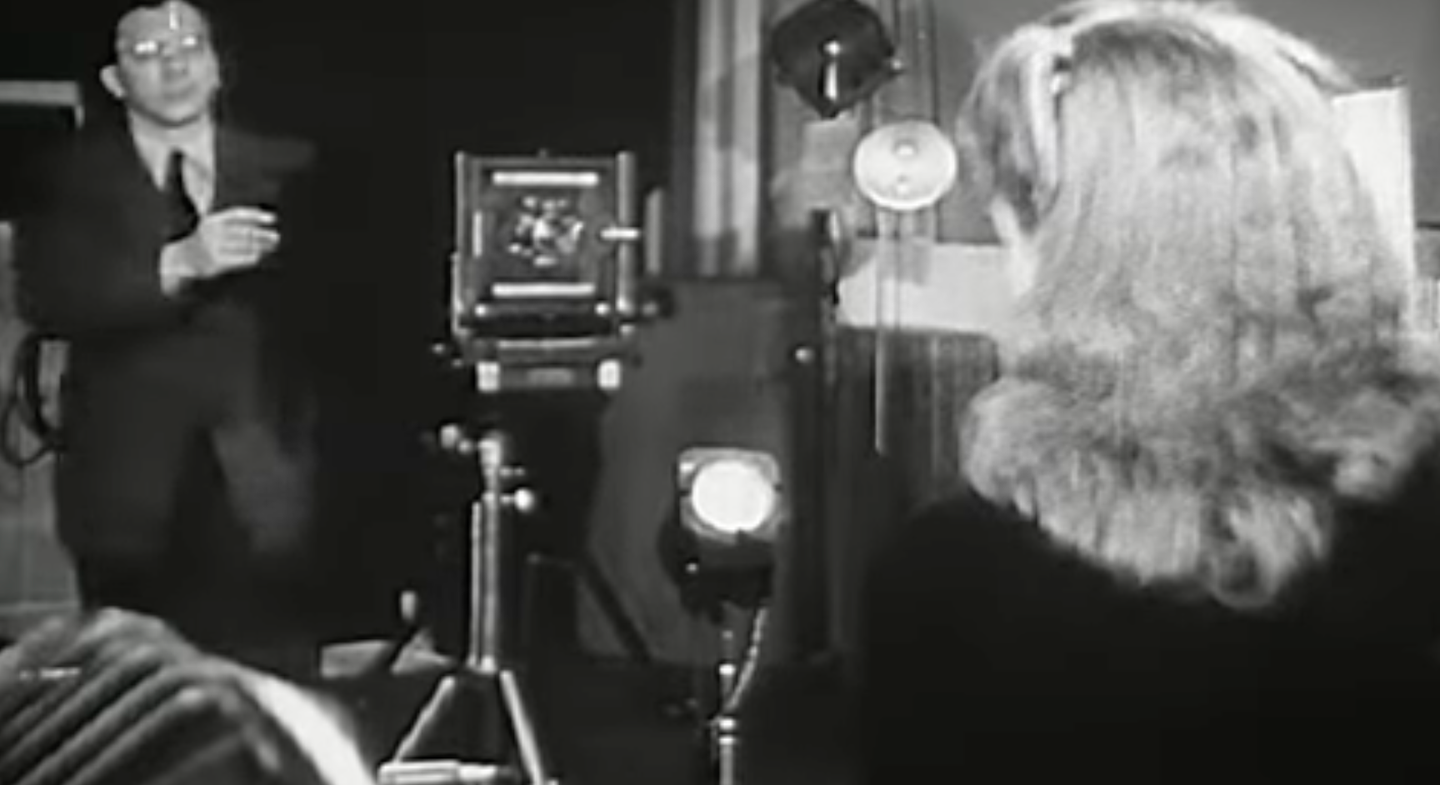 A still from a video about being a professional photographer in 1946. It shows a photographer standing next to a view camera with a model in the foreground.