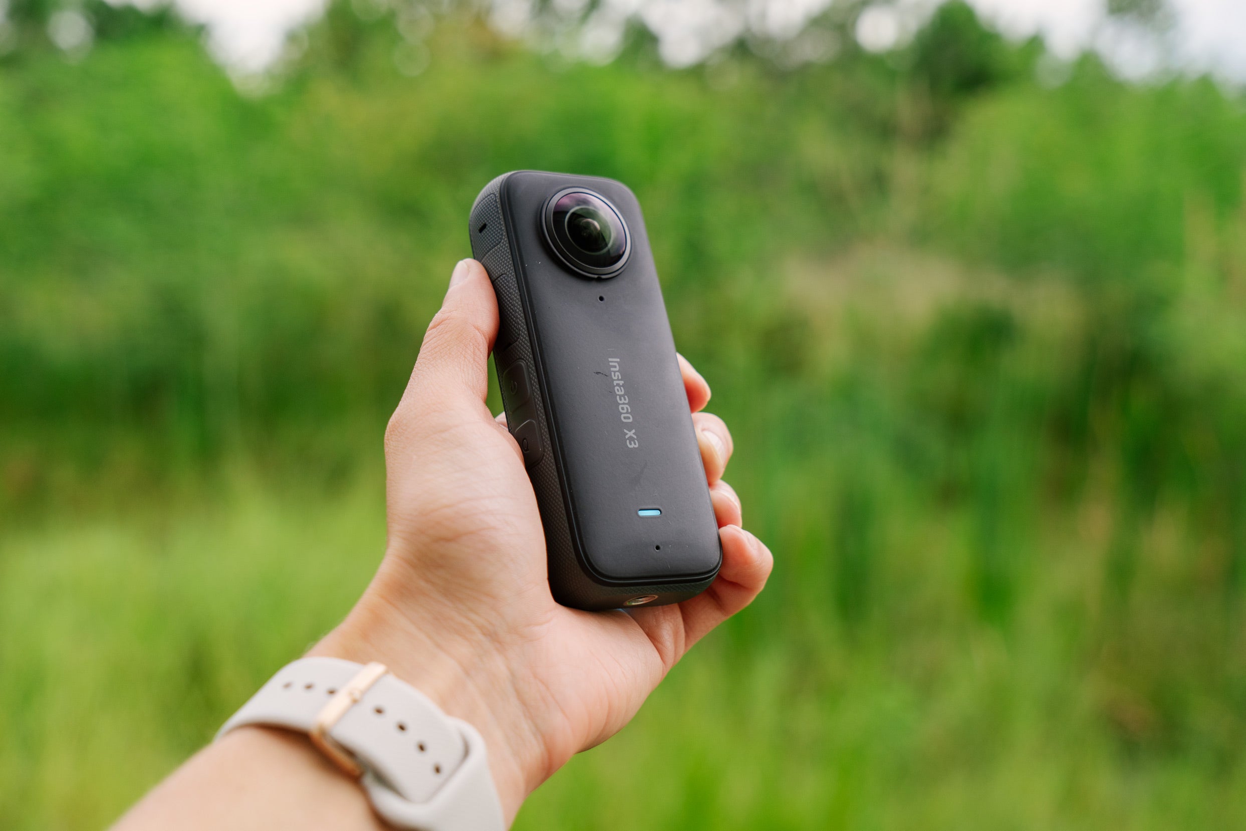 The Insta360 X3 held in a hand in front of a green forest