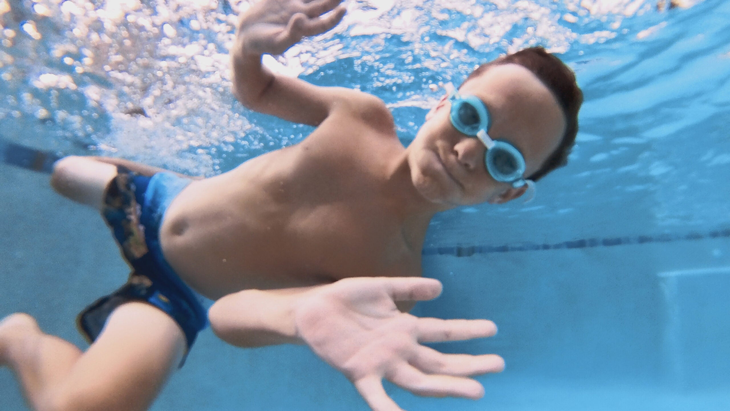 a kid swims underwater with goggles on, waving to the camera