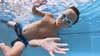 a kid swims underwater with goggles on, waving to the camera