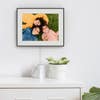 The Aura Walden digital photo frame with black frame displaying a family photo of three young children, hanging on a white wall.