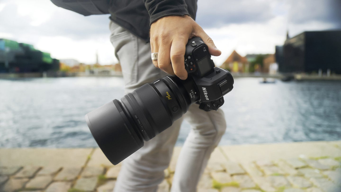 A hand holds a camera with Nikon NIKKOR 135mm f/1.8 S Plena lens attached.