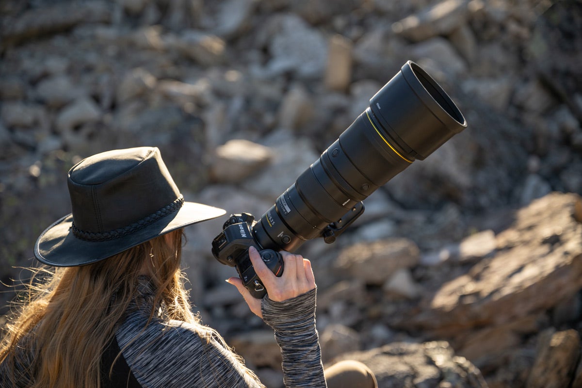 A woman holds a Nikon camera with the Nikon Z 600mm f/6.3 VR S lens attached with a rocky canyon in the background